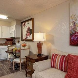 A cozy living room with a sofa, decorative pillows, and a table lamp, adjacent to an open kitchen and dining area adorned with artwork.