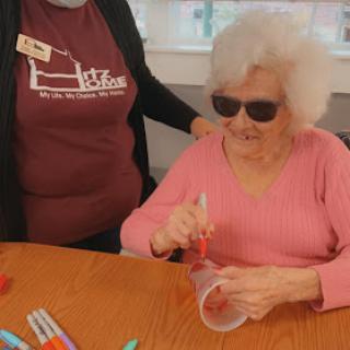 An elderly woman wearing sunglasses enjoys a crafting activity at a table, assisted by a caregiver.