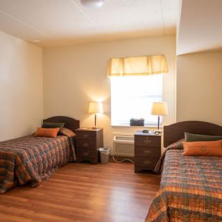 A cozy bedroom with two single beds, each adorned with decorative pillows, flanking a central nightstand under a softly glowing lamp. Natural light filters through the window, highlighting the warm wooden flooring.
