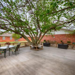 A spacious wooden deck with a large tree in the center, surrounded by outdoor seating and a brick wall backdrop.