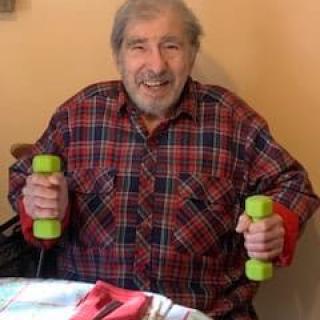 An elderly man smiles while holding dumbbells indoors, wearing a plaid shirt.