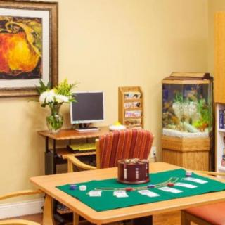 A cozy office space featuring a desk with a computer, a flower arrangement, a painting on the wall, an aquarium, and a table set up for card games.