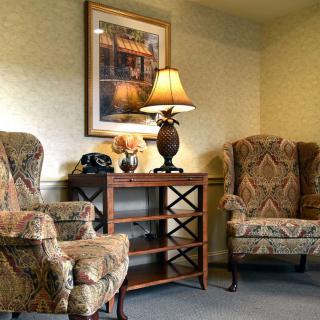 A cozy living room with two patterned armchairs, a wooden table with a lamp, and a framed painting on the wall.