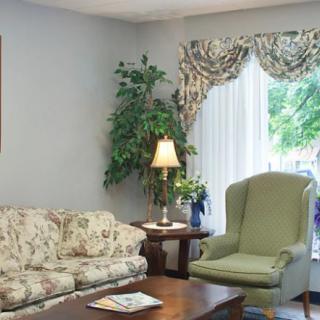 A cozy living room with a floral sofa, green armchair, side table with a lamp, and large window with curtains.