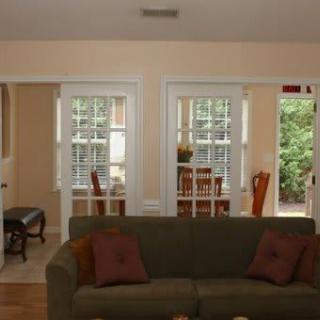 A cozy living room with a green couch facing toward the dining area, featuring large windows and warm lighting.