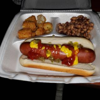 A meal consisting of a hot dog with ketchup and mustard, accompanied by tater tots and baked beans in a takeout container.