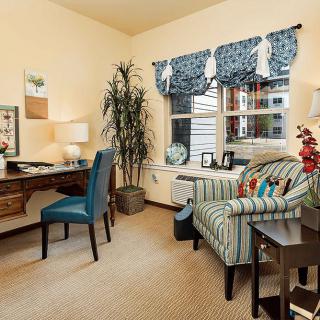 A cozy home office with a wooden desk and chair, decorative plants, and a comfortable armchair near the window.
