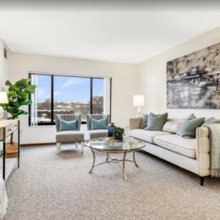 A spacious and elegantly decorated living room featuring a large sofa, coffee table, artwork, and ample natural light from a big window.