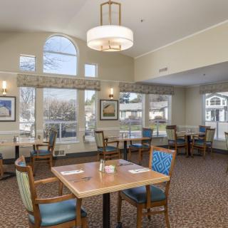 A well-lit dining area with multiple tables and chairs, featuring large windows and a chandelier.