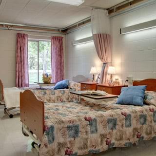A hospital room with two patient beds, decorated with patterned blankets and pillows, featuring a window with curtains.