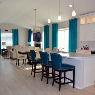 A modern open-plan kitchen and dining area featuring blue bar stools, pendant lighting, and large windows with blue curtains.