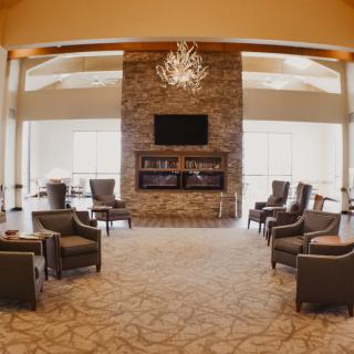 A spacious lounge with armchairs arranged around a central fireplace, adorned with a chandelier and featuring a television above the mantel.