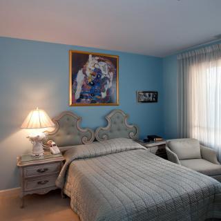 A cozy bedroom with blue walls, featuring a bed, nightstands, and a chair, adorned with a decorative painting.