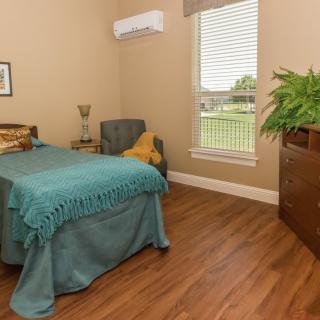 A cozy, minimalist bedroom featuring a single bed with teal bedding, wooden flooring, and a large plant near the window.