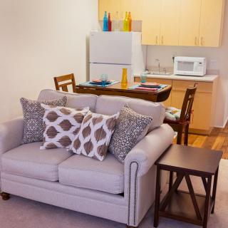 A cozy living area with a sofa and pillows near a small kitchen space equipped with a fridge and microwave.