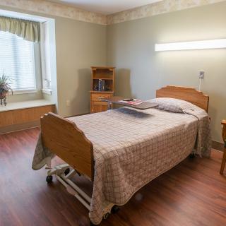 A clean and tidy hospital room with a single bed, wooden furniture, and natural light coming through the window.
