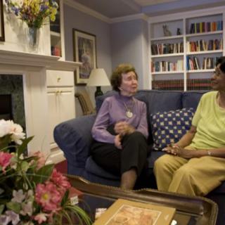 Two people having a conversation in a cozy living room with flowers and bookshelves.