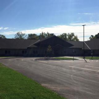 A one-story building with a large parking lot in front, surrounded by trees and clear blue sky.