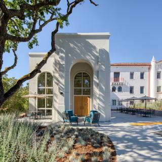 A modern building with arched windows and outdoor seating surrounded by trees under a clear blue sky.