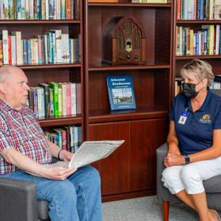 Two people sitting in a library engaged in conversation, with one person holding a newspaper.