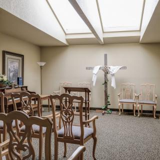 A small chapel interior with arranged chairs, a cross adorned with white drapery, and natural light streaming through skylights.
