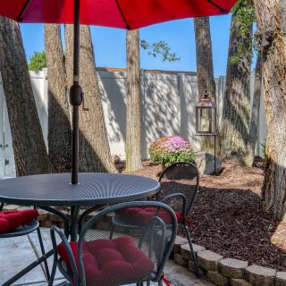 A cozy outdoor patio with a red umbrella and garden furniture, surrounded by trees and a landscaped yard.
