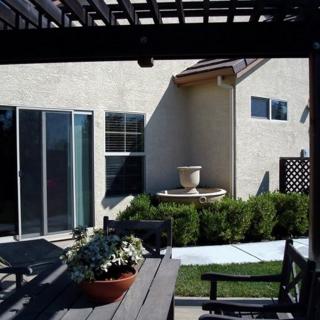 A cozy backyard patio with a pergola, table, chairs, and potted plants next to a house with a sliding glass door.
