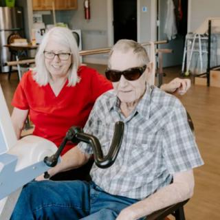 An elderly couple, with the man in a wheelchair, sits indoors smiling at the camera. He is wearing sunglasses.
