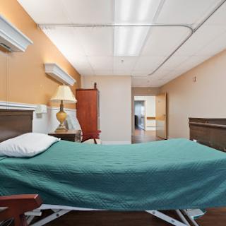 A well-lit hospital room featuring a bed with a green blanket and a bedside lamp.