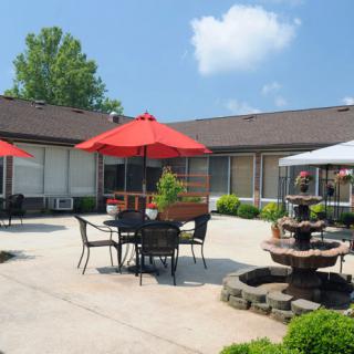 A sunny outdoor patio area with tables, chairs, red umbrellas, and a fountain.