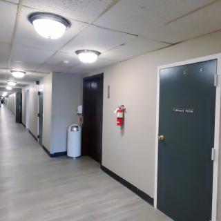 A hallway with several doors, ceiling lights, a fire extinguisher, and a trash can.
