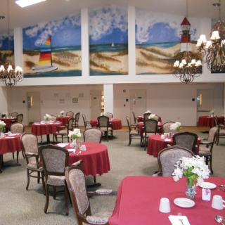 An elegant dining room with tables covered in red tablecloths, adorned with flower arrangements and set beneath large murals and chandeliers.