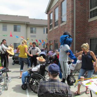 A lively outdoor event with people gathered, including a mascot in costume and individuals in wheelchairs, surrounded by festive decorations.