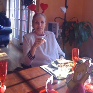 An elderly woman wearing a heart-shaped headband sits at a dining table with food and decorations.
