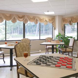A cozy recreation room with tables and checkers set up, featuring large windows and indoor plants.