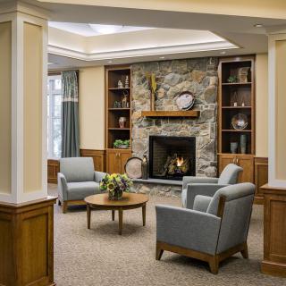 A cozy living room with a stone fireplace, surrounded by comfortable armchairs and wooden shelves.