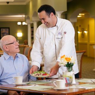 A chef serving a meal to a senior man in a dining room setting.