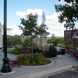 A small urban park featuring a statue surrounded by benches, trees, and landscaping.