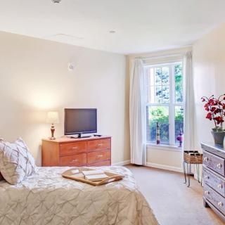 A cozy bedroom with a bed, dresser, and television, warmly lit by natural light from a large window.