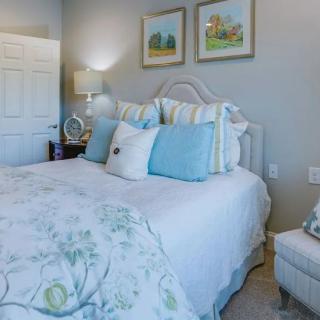 A cozy and elegantly decorated bedroom featuring a neatly made bed with blue and white pillows, a lamp on the bedside table, and framed artwork on the walls.
