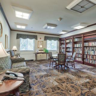 A cozy library room with bookshelves, a seating area, carpeted flooring, and soft lighting from lamps.