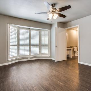 A room with hardwood floors, a ceiling fan, and window shutters, featuring a doorway leading to a bathroom.