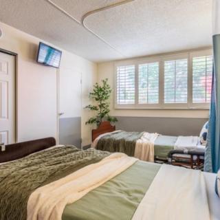 A well-lit hospital room with two neatly made beds, a wall-mounted television, and a large window with shutters.
