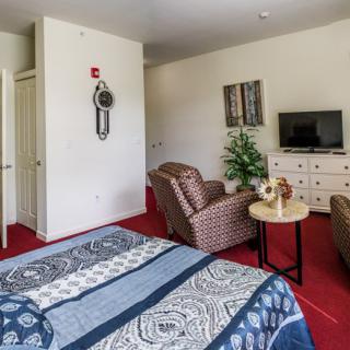 A cozy bedroom with a patterned bedspread, armchairs, a TV on a dresser, and red carpet flooring.