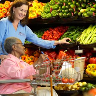 A caregiver assists an elderly woman in a vibrant grocery store filled with fresh fruits and vegetables.