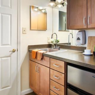 A compact kitchenette with wooden cabinets and a sleek countertop, featuring a sink and mirror.