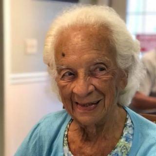 An elderly woman smiling warmly indoors, wearing a blue top.