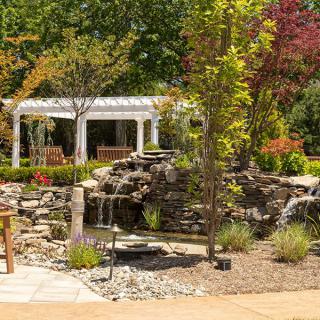 A serene garden featuring a waterfall, pergola, and outdoor seating surrounded by lush trees.