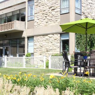 A modern building with a patio area featuring a table and chairs under a green umbrella, surrounded by a garden with flowers.