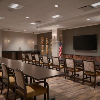 A well-lit conference room featuring a long table, multiple chairs, a large television on the wall, and an American flag in the corner.